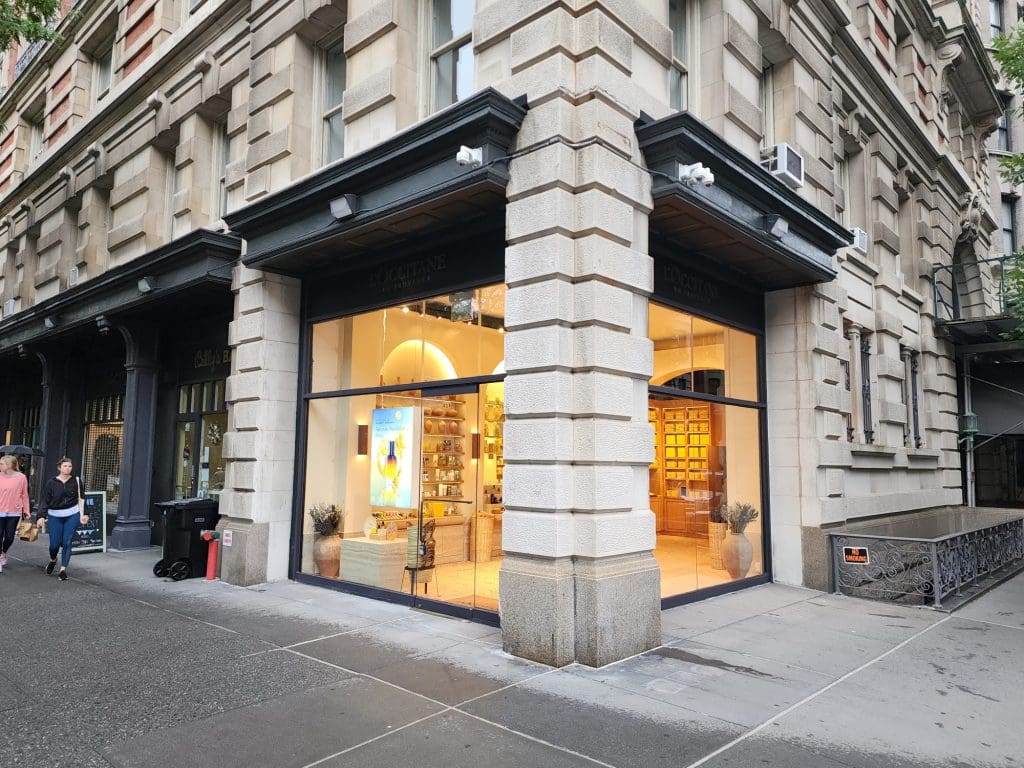 A vibrant street view of a corner storefront on Columbus Ave showcases large windows displaying various items inside. As part of the Columbus Ave Project, people stroll by on the bustling sidewalk, adding to the lively atmosphere.