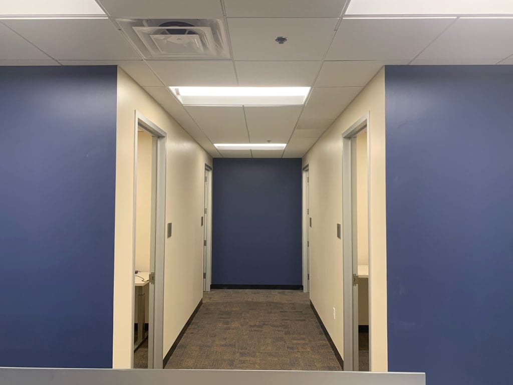 The office hallway, featuring blue and white walls, four doorways, and a carpeted floor, leads to the new focus room. Ceiling lights illuminate the space.