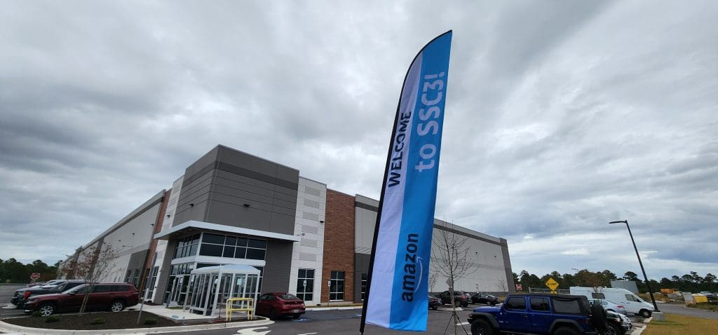 A modern warehouse with the Amazon logo prominently displayed and a "Welcome to SSC3!" banner flutters against a cloudy sky. Parked cars line the front, hinting at the bustling activity inside.