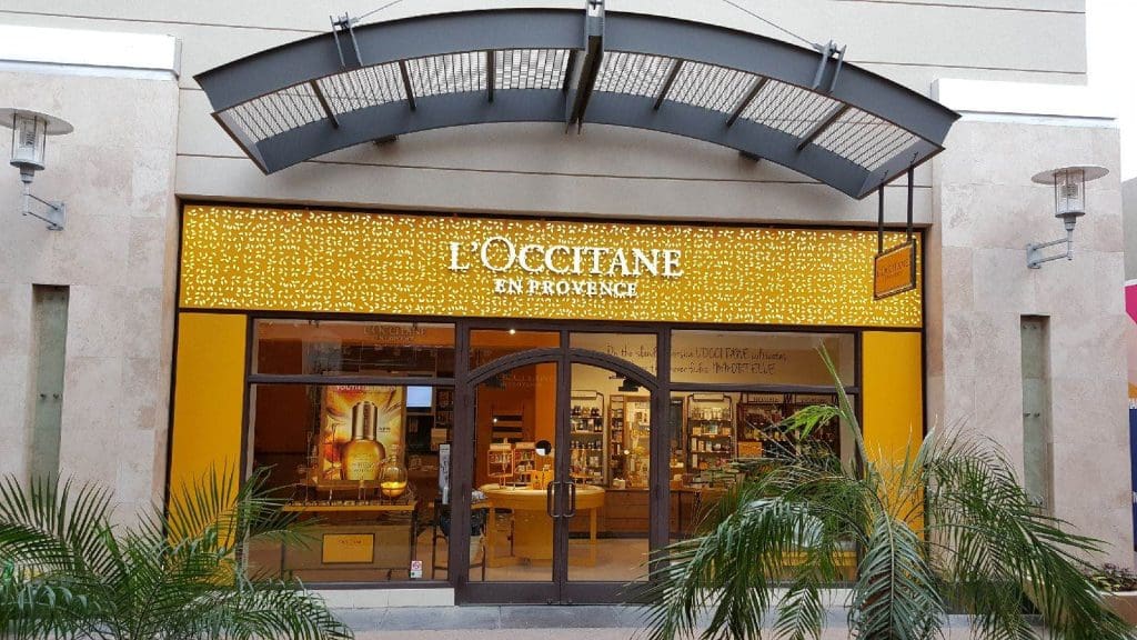 The storefront of L'Occitane en Provence at Biltmore Fashion Park features a lit sign, large glass doors, and lush tropical plants in front.