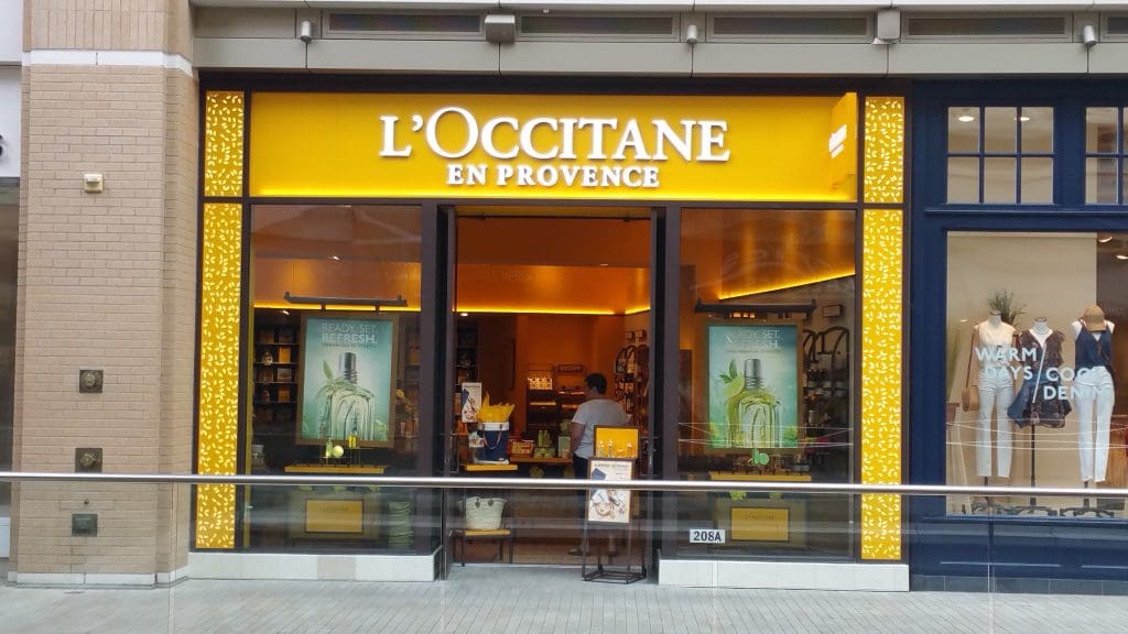 Front view of a L'Occitane en Provence store at City Creek Center, with a bright yellow sign and large promotional posters on either side of the entrance.