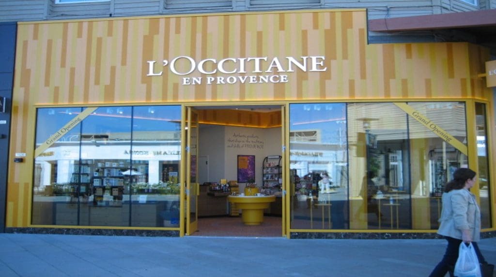 The storefront of L'Occitane en Provence at the Village at Corte Madera boasts a vibrant yellow and orange exterior. A woman strolls past on the sidewalk, enjoying a leisurely day.