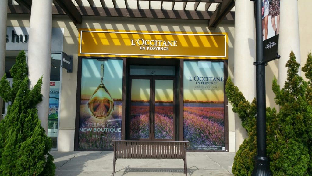 The storefront of L'Occitane en Provence at St John's town center features large ad images, with a welcoming bench in front.