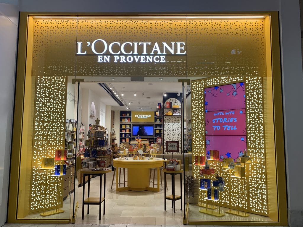 Front view of a L'Occitane en Provence store at Lennox Square, with illuminated signage and decorative facade. A display of various products on shelves and tables is visible through the glass entrance, inviting shoppers inside this vibrant shopping mall.