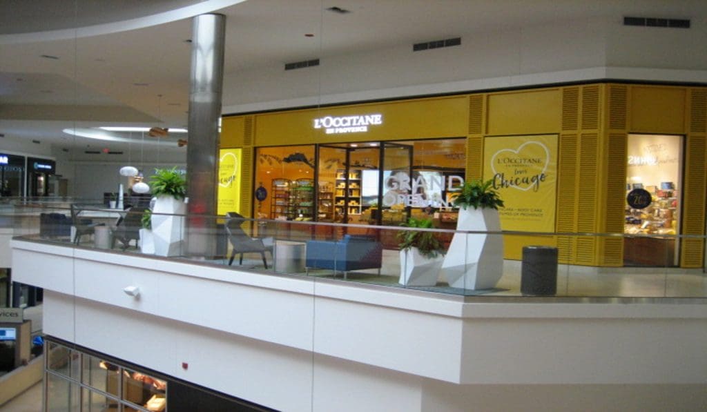 The storefront of L'Occitane en Provence at the Chicago Fashion Outlets showcases its bright yellow signage and "Grand Opening" text. Surrounded by sleek, modern planter decorations and a glass railing on the upper level, it exudes an inviting ambiance in this bustling retail destination.