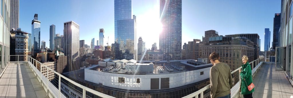 Panoramic view of a city skyline with tall buildings, a bright sun, and two people on a balcony enjoying their new home in the foreground.