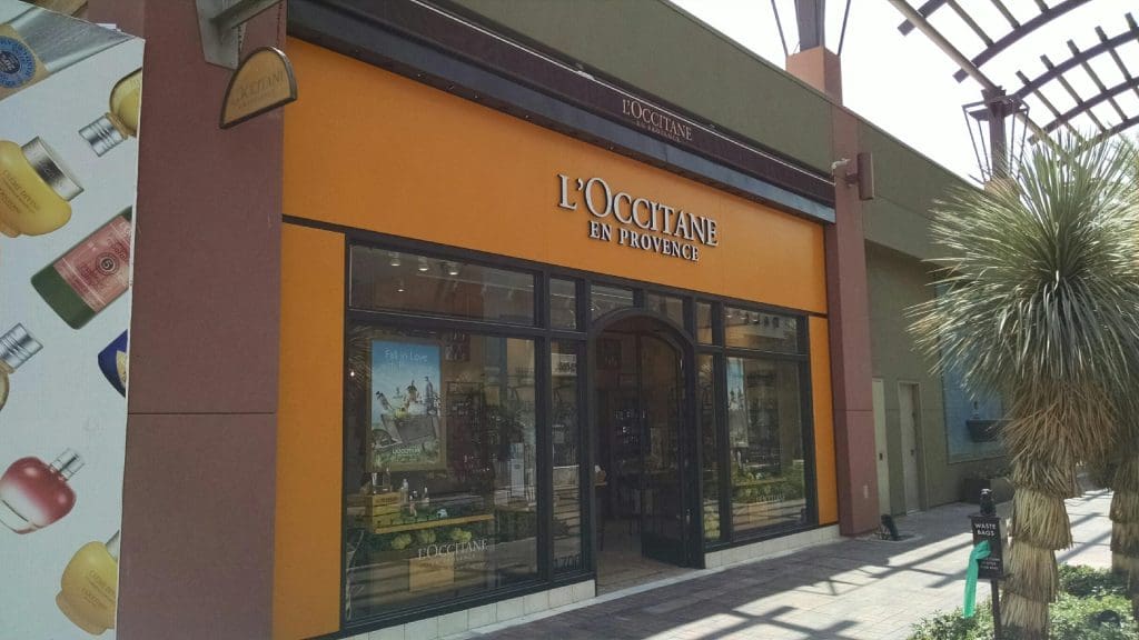 The storefront of L'Occitane en Provence at El Paseo Gardens features orange accents and glass doors, showcasing products inside. Nearby, a palm tree sways gently next to an eye-catching advertisement.