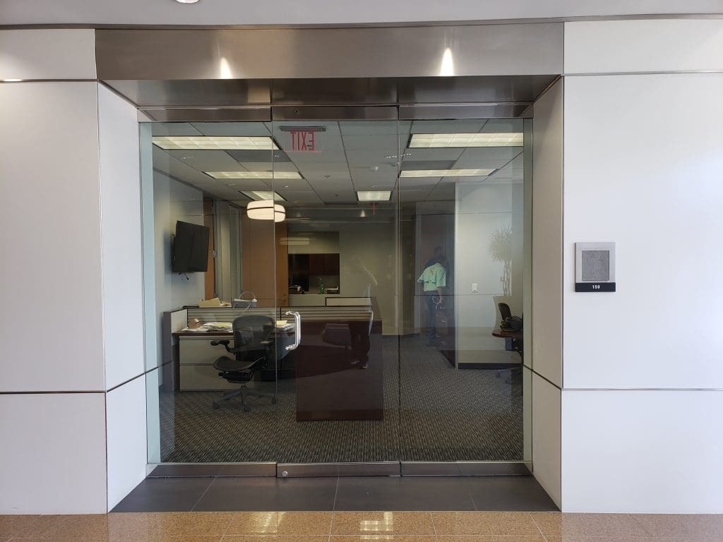 Office entrance with glass doors, giving a glimpse into an advisor's domain with neatly arranged desks and chairs. An exit sign is prominently visible above the door, ensuring clear guidance for all within the office program's lively environment.