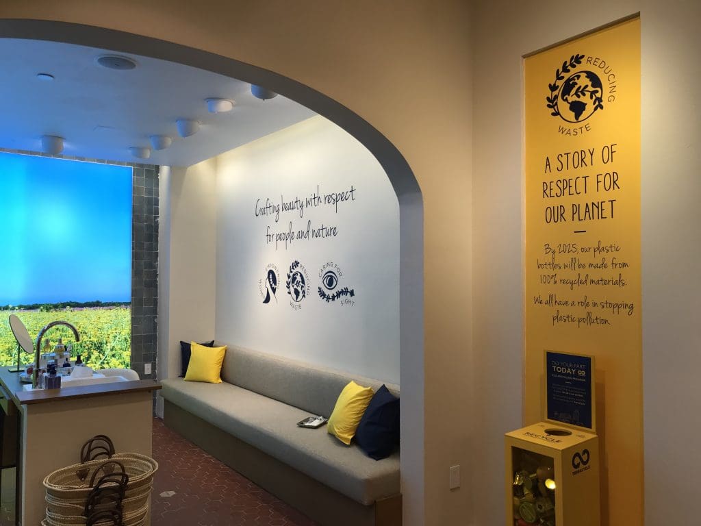 Nestled within Westfarms Mall, this cozy store interior features a seating area and eco-friendly messaging on the walls. A display promotes reducing waste, aiming for plastic replacement by 2025. Natural light streams through a large window, creating a warm retail store ambiance.