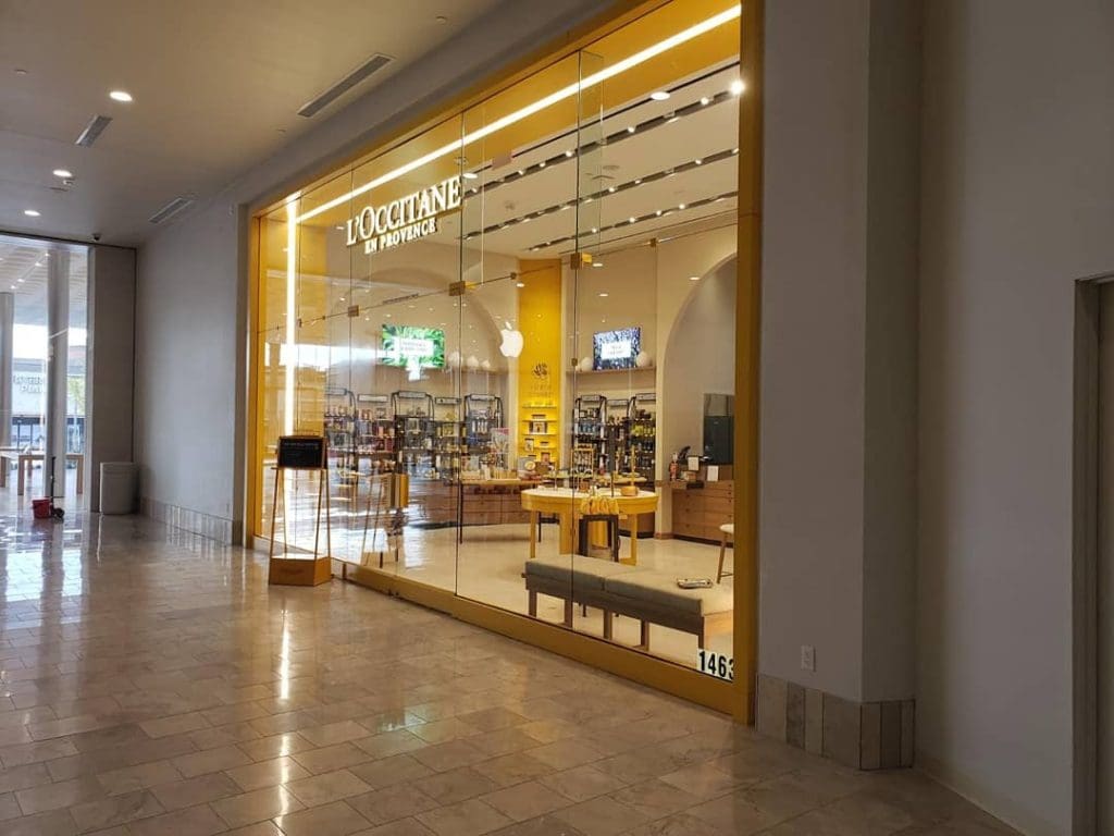 The storefront of L'Occitane en Provence in Scottsdale features a bright yellow frame, perfectly complementing the vibrant fashion district of the square. Through the display window, you can glimpse an inviting array of products and well-organized shelves inside.