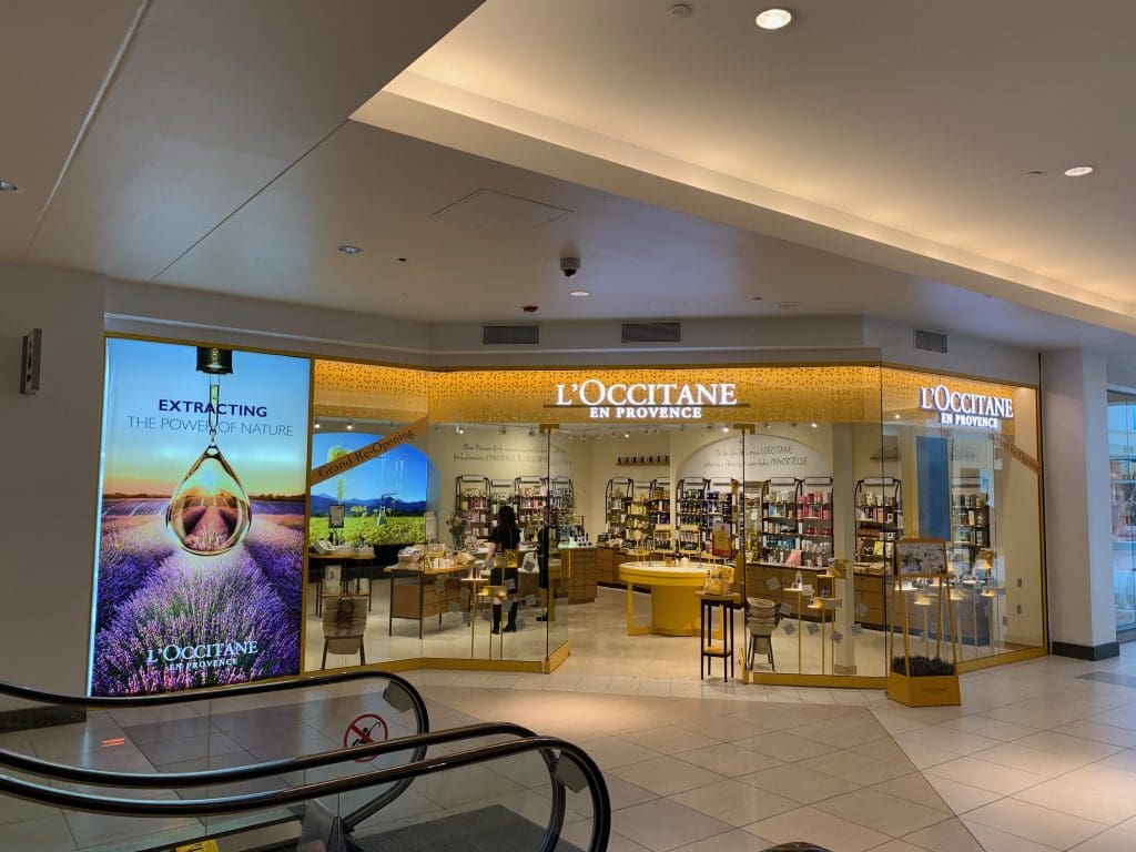At the Fashion Mall, the L'Occitane en Provence store boasts illuminated signage and exquisite product displays visible through its glass storefront. An escalator glides nearby, crafting a seamless shopping experience.