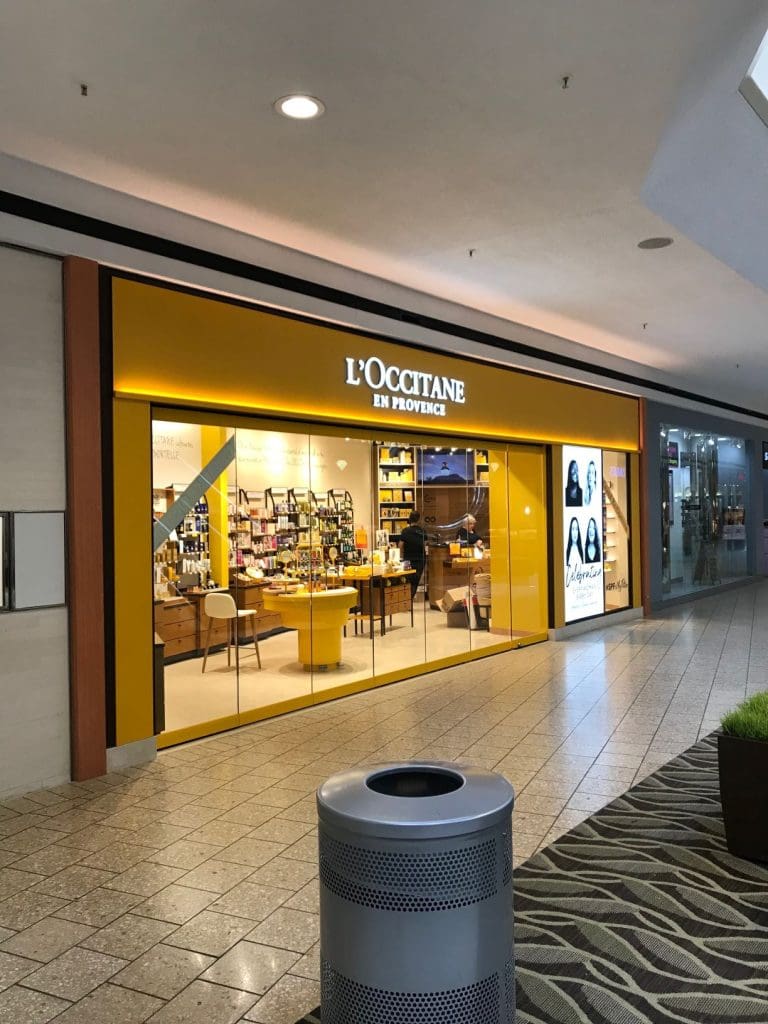 The storefront of L'Occitane en Provence in Fair Oaks Mall features its signature yellow signage and display windows showcasing an array of luxurious products. A trash bin is subtly positioned in the foreground, adding a touch of practicality to this elegant shopping destination.
