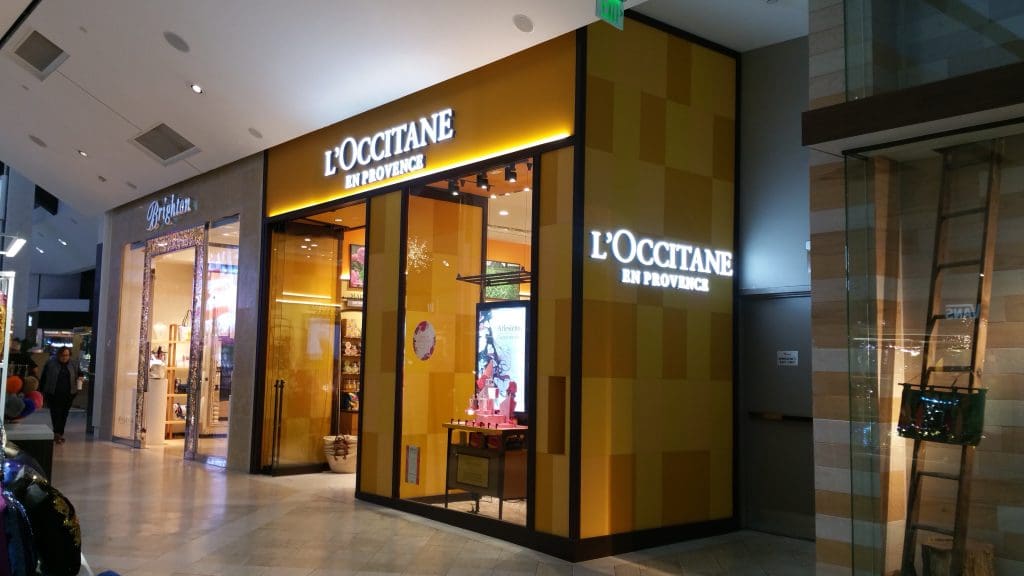 The storefront of L'Occitane en Provence in the shopping mall boasts a fashion-forward flair with its vibrant yellow exterior and illuminated signage, perfectly complementing the bustling energy of the square.