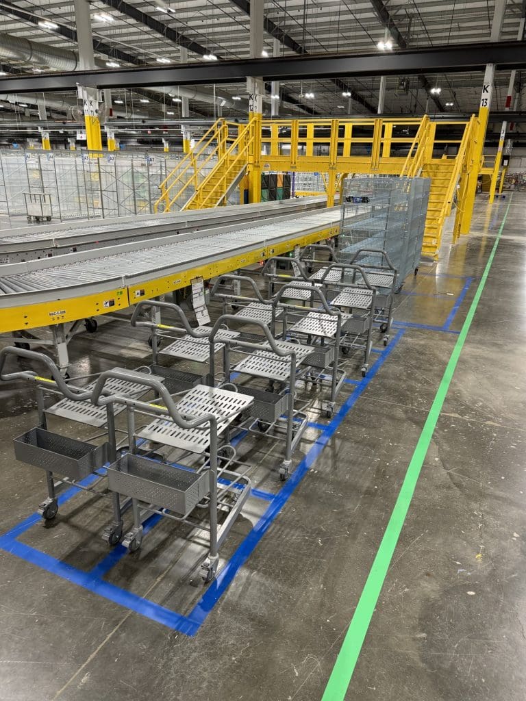 The warehouse interior features yellow stairs, bustling conveyor belts, and a row of metal carts neatly lined up on a marked floor path, efficiently managed by the RSR+ system.