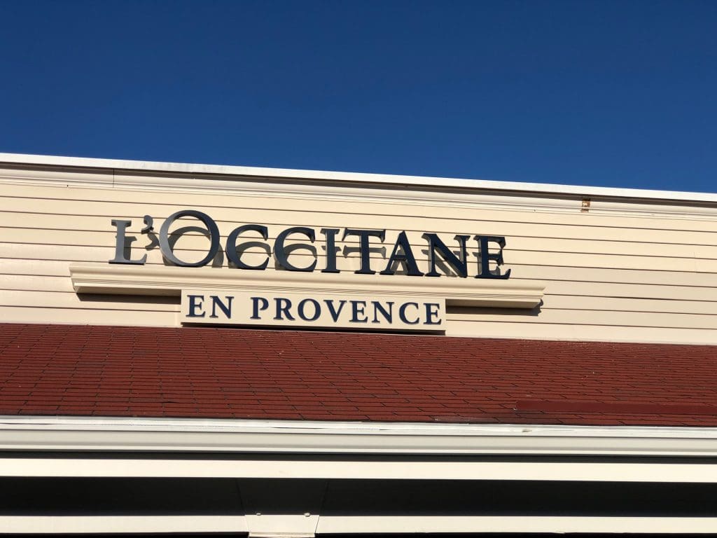 A L'Occitane en Provence outlet stands proudly with its cream-colored facade and red roof, nestled under a clear blue sky in Wrentham, inviting all who pass by to indulge in a delightful shopping experience.