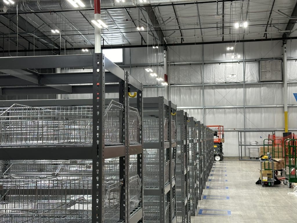 A large warehouse interior with empty metal shelving units lined up in rows, illuminated by visible overhead lighting, provides an ideal space for the RSR+ PROGRAM.