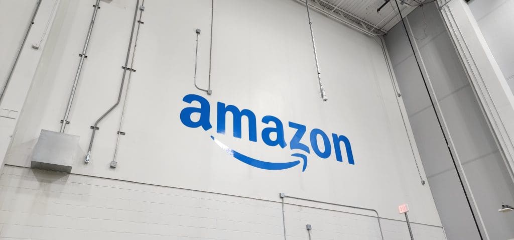 A large Amazon logo adorns a white wall inside the SFL7 warehouse, characterized by its high ceilings and visible electrical conduits.
