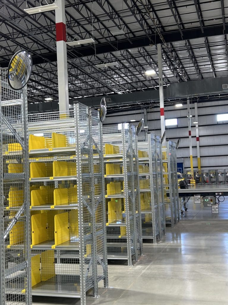 The warehouse interior, part of our RSR+ Program, features metal shelving units—some empty and others housing yellow bins. The high ceilings with metal beams and a conveyor system in the background highlight its efficient design.