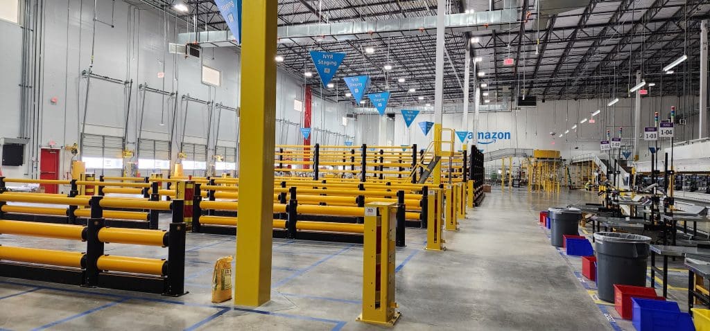 The interior of the large warehouse features yellow safety barriers, shelves stacked neatly, and blue banners displaying the latest SSD program. Various equipment is arranged methodically in this well-lit and organized space, ensuring efficient operations.