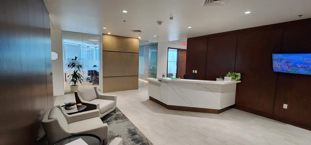 The modern office reception area features a sleek white desk, chairs, and lush plants, creating a refreshing atmosphere. A wall-mounted screen enhances the space, while frosted glass walls offer a glimpse into the conference room—a perfect setting for any wealth advisor meeting.