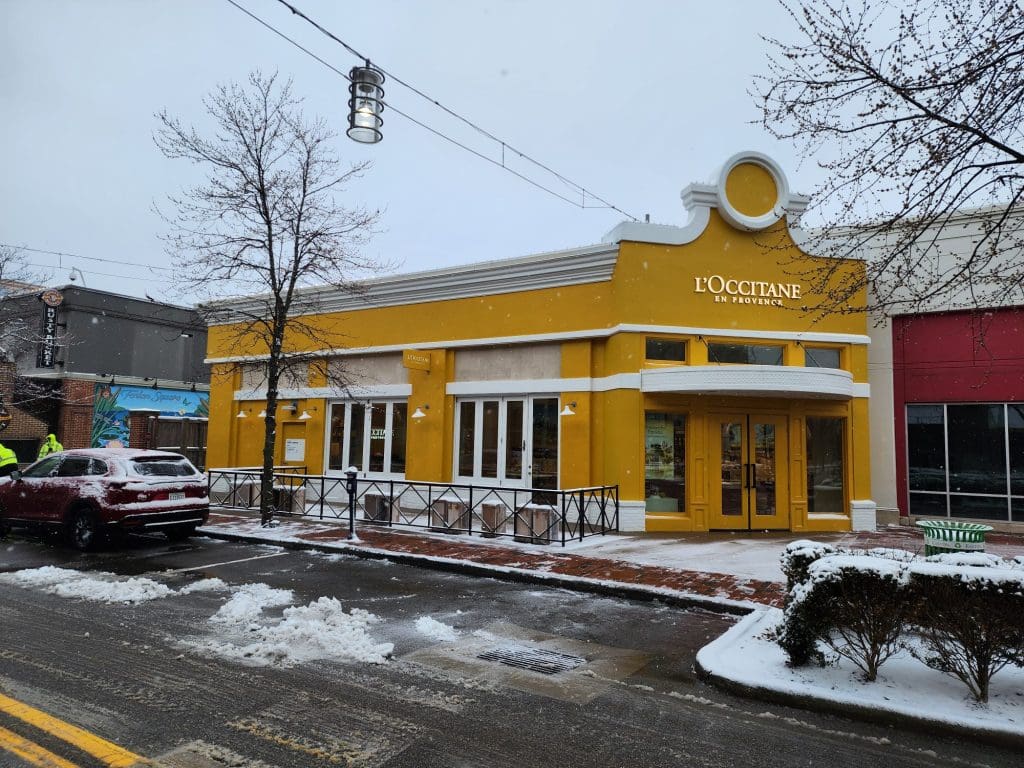 The yellow storefront of L'Occitane en Provence brightens a snowy Columbus street near Easton Town Center, with a parked car and bare trees adding to the quintessential shopping scene.