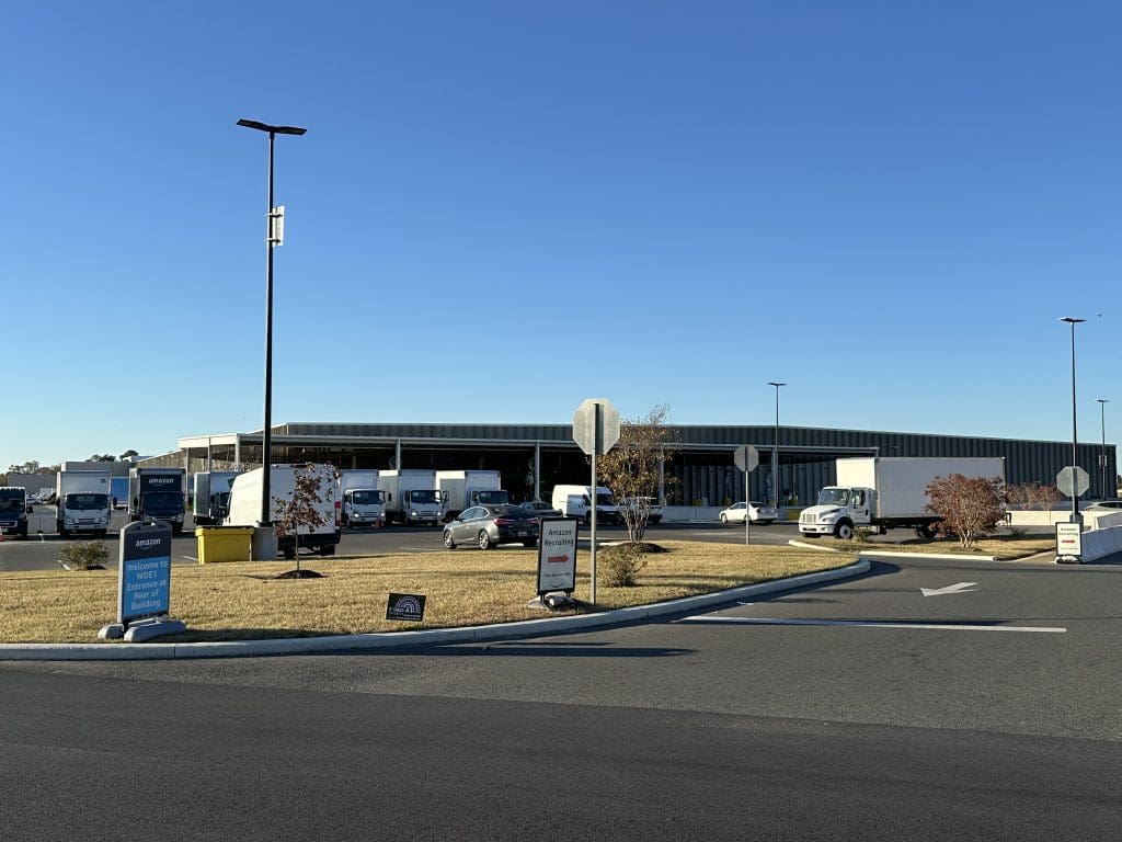 A fleet of white trucks, part of the RSR+ PROGRAM, is parked outside a large warehouse under a clear blue sky. Signs and a circular road add structure to the foreground scene.