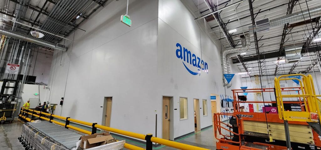The interior of an Amazon warehouse buzzes with activity as conveyor belts whir alongside shelves stocked with SSDs. A large wall displays the iconic Amazon logo, surrounded by various equipment and safety signs, while a SAX5 program ensures efficiency and precision in every operation.