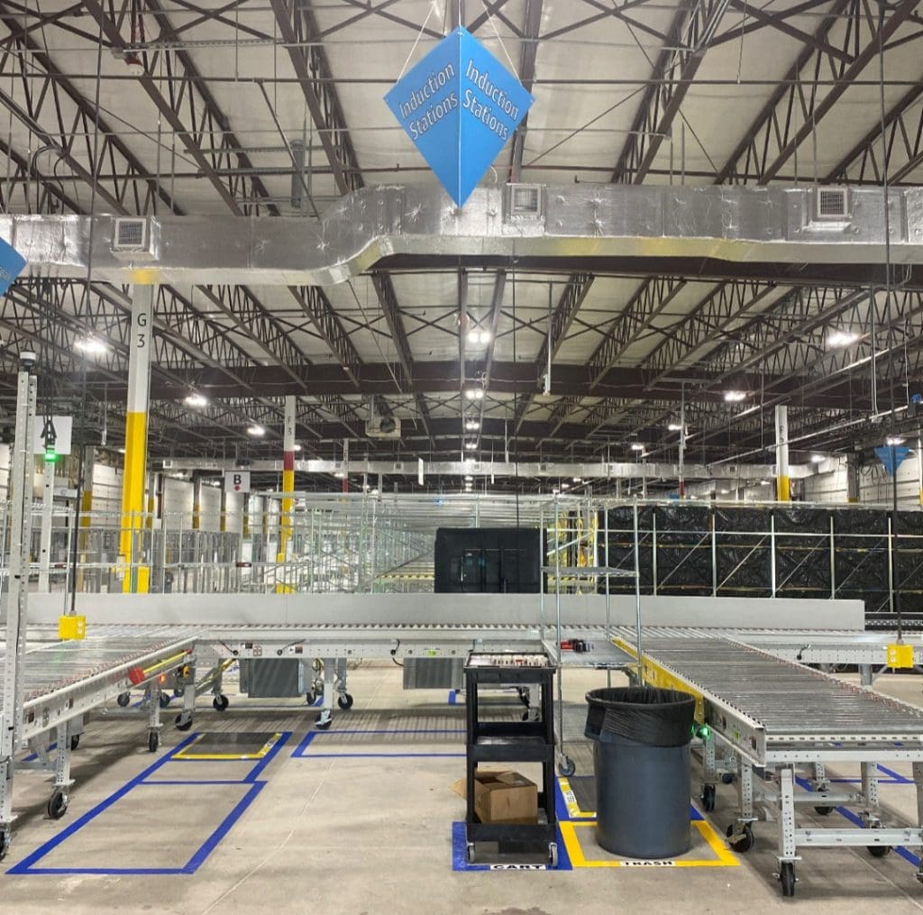 Warehouse interior with empty conveyor belts and metal racks under the industrial ceiling, labeled as "Induction Stations" for WNB2.