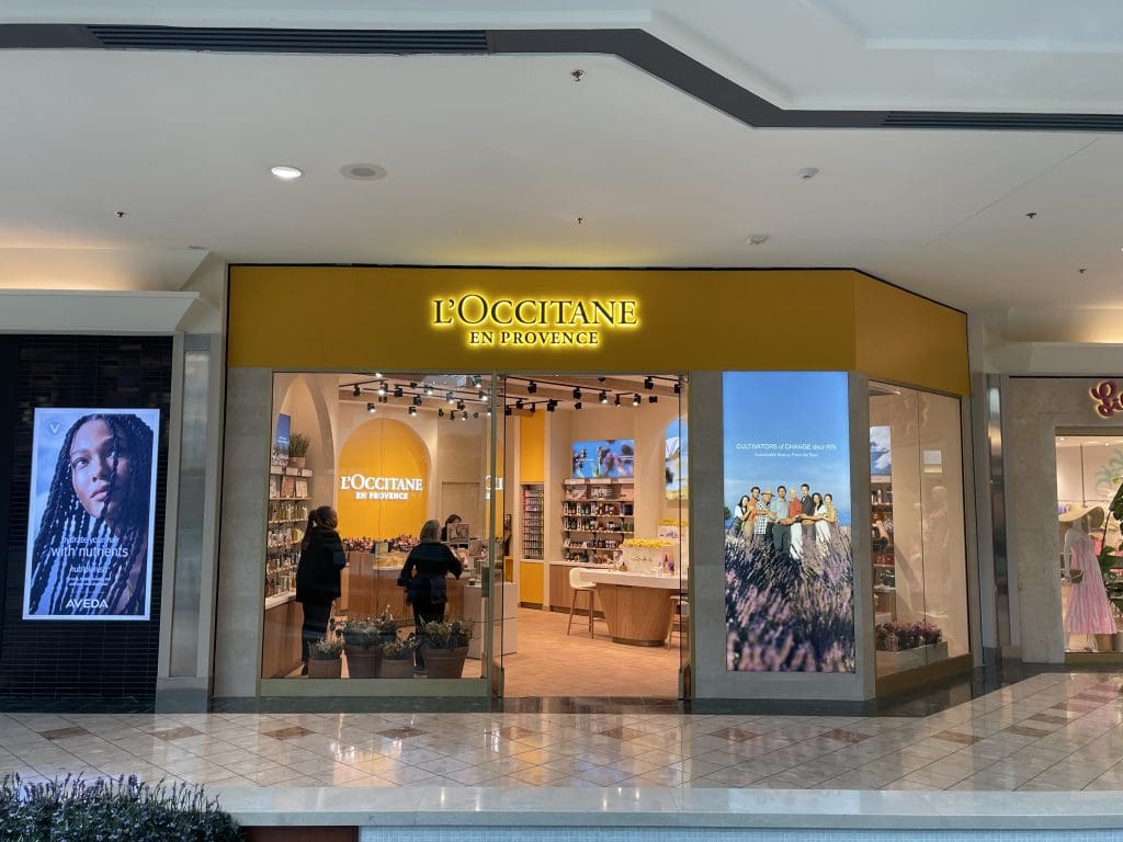 The storefront of L'OCCITANE en Provence in the Gardens Mall features two customers immersed in their shopping experience. Large promotional posters adorn both sides of the entrance, showcasing the brand's retail allure.