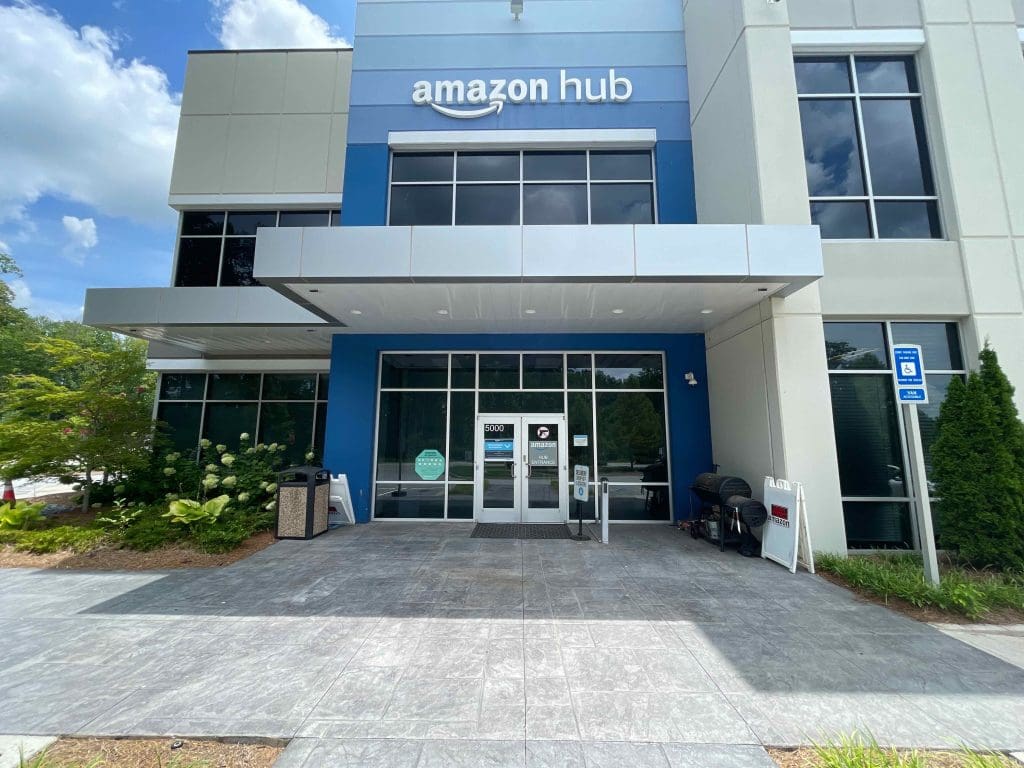 The exterior of the Amazon Hub building, featuring a PROGRAM Locker+ system, boasts a glass entrance with blue accents and is enhanced by surrounding plants under a clear sky.